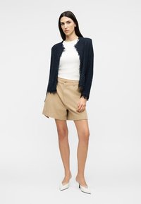 Γυναίκα με υφασμάτινο μπλε σκούρο σακάκι, λευκό τοπ, μπεζ σορτς και λευκά μυτερά ψηλοτάκουνα, στέκεται μπροστά σε απλό φωτεινό φόντο.