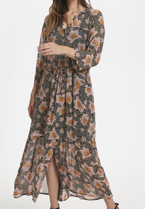 Femme portant une robe à manches longues et col en V, à motifs floraux roses et orange sur fond sombre, avec une fente à l'avant et une ceinture à la taille.