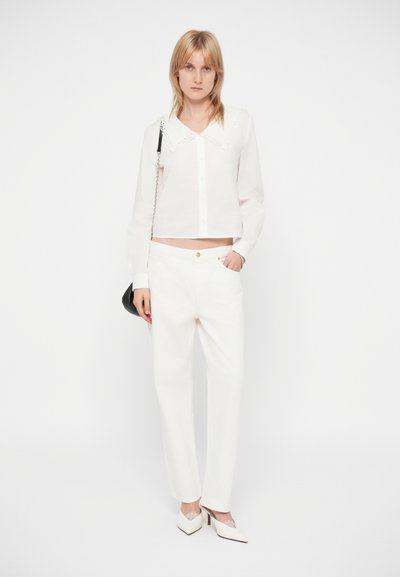 Femme avec des cheveux blonds raides portant un chemisier blanc à manches longues avec un col en dentelle, un pantalon blanc, des talons blancs et un sac à épaule noir.