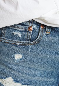 Wyblakłe niebieskie dżinsy z postarzanymi detalami i miedzianymi guzikami, charakteryzujące się postrzępioną teksturą i klasycznym pięciopocketowym krojem.