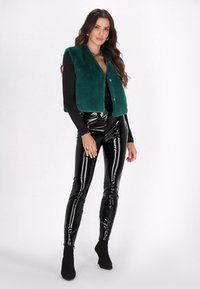 Vesta scurtă din blană sintetică teal, cu guler tip șal și nasturi aurii, purtată cu pantaloni mulați negri lucioși și botine negre.