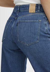 Jeans de mezclilla azul de talle alto con un acabado texturizado, dos bolsillos traseros y una etiqueta de marca de cuero marrón en la cinturilla.