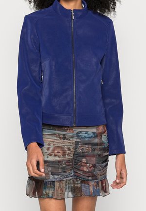 Woman wearing a royal blue textured zip-up jacket and a gathered mini skirt with a multicolored abstract print and ruffled hem.