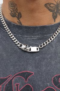 Zilveren ketting met een doodshoofdsieraad en een sluiting, gedragen over een grijze shirt met een vervaagd grafisch ontwerp.