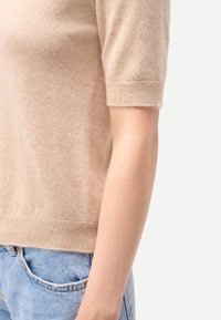 Pull beige à manches courtes avec une texture douce et un ourlet côtelé, porté avec un jean en denim bleu clair. Vue en gros plan de côté.