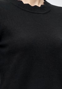 Gros plan d’une personne portant un pull en maille noire avec un col rond légèrement froncé sur un fond uni.