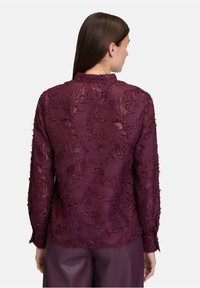 Donna con capelli castani lisci che indossa una blusa lunga a maniche corte in bordeaux trasparente e testurizzata e pantaloni larghi abbinati, di spalle.
