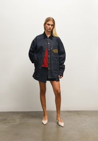 Mørkeblå denimjakke med krage, frontlommer og brodert "HOUSE OF SUNNY"-logo. Båret over en rød genser og matchende denimskjørt.