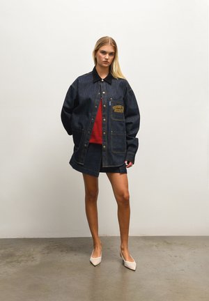 Giacca di jeans blu scuro con colletto, tasche frontali e logo ricamato "HOUSE OF SUNNY". Indossata sopra un maglione rosso e una gonna di jeans abbinata.