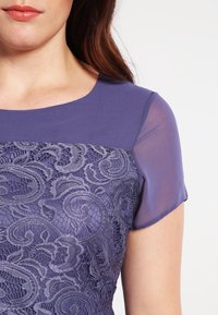 Robe violette avec un corsage en dentelle texturée ornée de motifs complexes, complétée par un tissu uni et lisse aux épaules et aux manches.