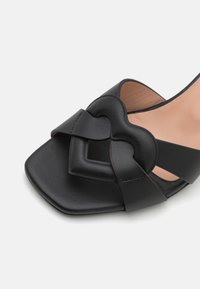 Melnas ādas sandalītes ar kvadrātveida purngalu, kurām ir sirds formas dizaina elements augšpusē un gluda, matēta apdare.