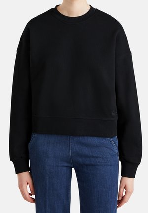 Person trägt einen schlichten schwarzen Sweatshirt mit Rundhalsausschnitt und langen Ärmeln und blaue Jeans, steht vor einem weißen Hintergrund.