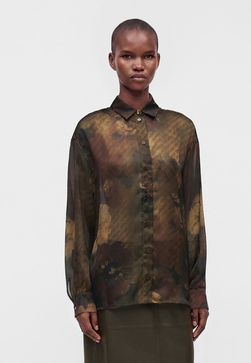 Femme portant un chemisier transparent à manches longues avec un motif floral foncé et un col, debout devant un fond uni.