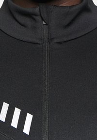 Schwarzes Sportoberteil mit hohem Kragen, verfügt über einen zentralen Reißverschluss und vier weiße Streifen nahe dem Saum, hergestellt aus glattem, dehnbarem Stoff.