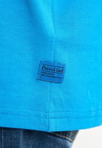 Světlá modrá tričko vyrobené z měkké bavlny, s obdélníkovou modrou visačkou, na které je nápis "Petrol Ind" a další text. Čisté šití na lemu.