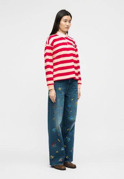 Γυναίκα όρθια, φορώντας κόκκινο και ροζ ριγέ μακρυμάνικο πόλο πουκάμισο, φαρδιά τζιν με κεντήματα λουλουδιών και καφέ παπούτσια σε απλό φόντο.