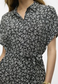 Vestito camicia nero con motivo floreale bianco, maniche corte, colletto e dettaglio in vita legato. Tessuto leggero con una superficie liscia.