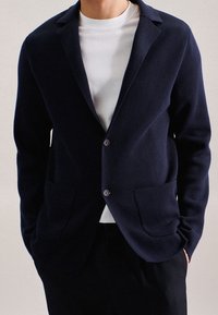 Mörkblå stickad cardigan med slagkrage, framfickor och två knappar. Lager på en vit t-shirt med rund hals, som har en struktur. 