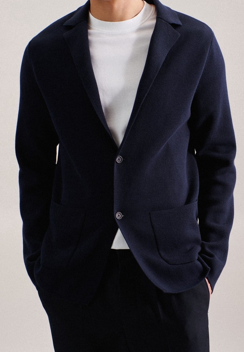 Mörkblå stickad cardigan med slagkrage, framfickor och två knappar. Lager på en vit t-shirt med rund hals, som har en struktur.