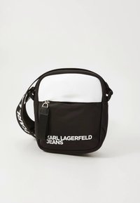 Černá kabelka přes rameno s bílým detailem nahoře. Má přední kapsu na zip, nastavitelný popruh a výrazné potištěné logo.