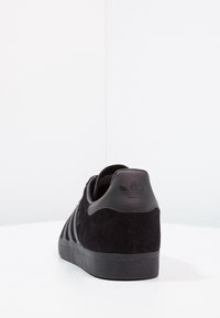 Baskets en daim noir avec trois bandes texturées, semelle en caoutchouc et une languette de talon noire mate avec un logo embossé. Design simple et moderne.