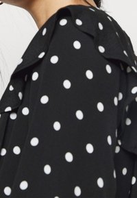 Blusa in tessuto nero con pois bianchi, dettaglio del colletto gessato e una texture morbida, parzialmente visibile in una vista ravvicinata.