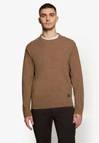 Brun uldtrøje med rund hals, ribbede manchetter og nedre kant, med et lille mærke på den nederste side. Båret med mørke bukser.