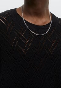 Zwarte gebreide trui met een gestructureerd chevronpatroon en een ronde halslijn, gedragen met een zilveren ketting rond de nek.