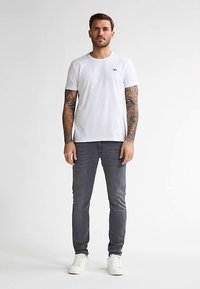 Biała koszulka z krótkim rękawem z małym logo, zestawiona z szarymi, dopasowanymi jeansami i białymi sneakersami. Na ramionach modela widoczne są tatuaże.