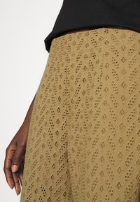 Χακί φούστα με περίπλοκα σχέδια σαν δαντέλα, σε χαλαρή γραμμή και υφή. Ορατό χέρι που στηρίζεται στην άκρη του υφάσματος.