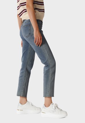 Persona che indossa jeans azzurri corti e sneaker bianche con dettagli argentati, in piedi davanti a uno sfondo semplice.