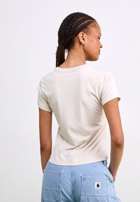 T-shirt beige à manches courtes avec un col rond, en coton. Le tissu a une texture lisse, avec des détails de couture visibles sur les manches.
