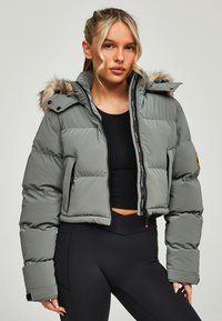 Korte grijze pufferjack met een hoed met bontafwerking, een rits aan de voorkant en elastische manchetten. Heeft zwarte logo-details langs de rits.