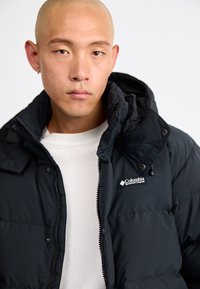 Puffjacka i mörk svart, med zip-stängning, huva och en logotyp på bröstet. Slät textur och quiltat mönster med modern passform.