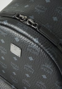 Zwarte leren rugzak met textuur, voorzien van een ritssluiting, een metalen logo plaat en een subtiel geometrisch patroon van grijze diamanten.