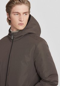 Giacca con cappuccio di colore marrone scuro realizzata in materiale liscio e resistente all'acqua. Presenta una chiusura con zip e un logo discreto sulla manica.
