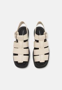 Beige sandalen met banden en zwarte zool, met een geweven ontwerp, open achterkant en verstelbare enkelband met gespsluiting.