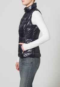 Mujer con chaleco sin mangas acolchado azul sobre una camisa de manga larga blanca y vaqueros azules, con la mano en el bolsillo, mirando hacia la derecha en un fondo neutro.