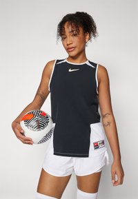 Czarny bezrękawnik sportowy z białymi akcentami, logo Nike, w połączeniu z białymi szortami. Trzyma wzorzystą piłkę nożną.