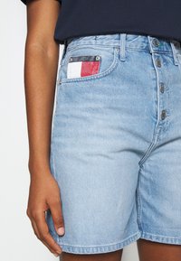 Shorts de mezclilla azul claro con cierre de cinco botones, un bolsillo frontal con un parche a cuadros en rojo, blanco y azul, y una tela texturizada.