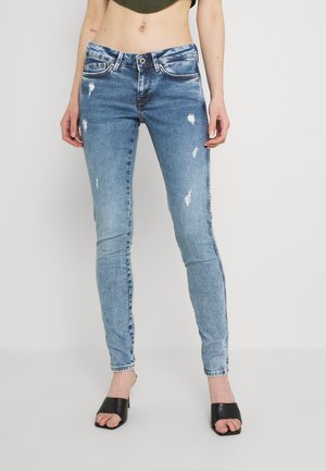 Slim fit lichtblauwe distressed jeans gedragen met zwarte open teen slip-on hakken, cropped view met zicht op de buik en benen tegen een effen achtergrond.