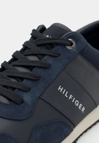 Granatowy sneaker wykonany z zamszu i skóry, z czarnymi sznurowadłami, metalowymi oczkami i białym logo po boku. Teksturowane akcenty.