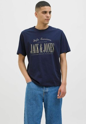 Marineblaue Baumwoll-T-Shirt mit kurzen Ärmeln und beigen Grafiken. Kombiniert mit hellblauen, locker sitzenden Jeans.
