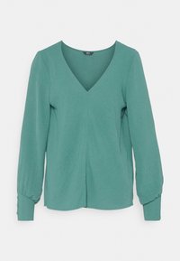 Modrá dlouhá halenka s výstřihem do V, nařasenými manžetami a knoflíkovými detaily na okrajích manžet, vystavená na bílém pozadí.