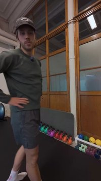 Atletisk mand i en grøn tekstureret sweater og grå shorts, stående ved siden af et stativ med farverige kettlebells på en sort gulv.