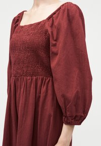 Γυναίκα που φοράει μακρυμάνικο μπορντό φόρεμα με τετράγωνο λαιμόκοψη σμάκι και φουσκωτά μανίκια που συγκεντρώνονται σε ελαστικές μανσέτες.