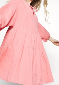 Rosa, gestuftes Kleid aus strukturiertem Stoff mit einem Schlüssel-ausschnitt und Bindeschluss. Verfügt über lange Ärmel und eine lockere Passform.