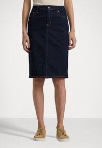 Jupe A-line en denim foncé avec une fente sur le devant, ornée de coutures orange et d'une fermeture à boutons classique. Associée à des chaussures à lacets beiges.