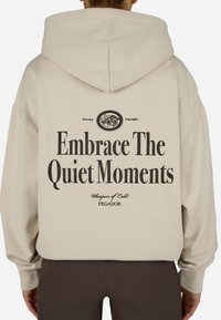 Mikina s kapucňou z béžovej tkaniny s uvoľneným strihom. Na chrbte sa nachádza výrazný čierny nápis: "Prijmi tiché chvíle" a grafika.