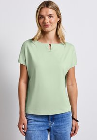 Světlá zelená krátká rukávová blůza s kulatým výstřihem a otvorem v límci. Vyrobena z měkké, hladké tkaniny. Klasický střih.